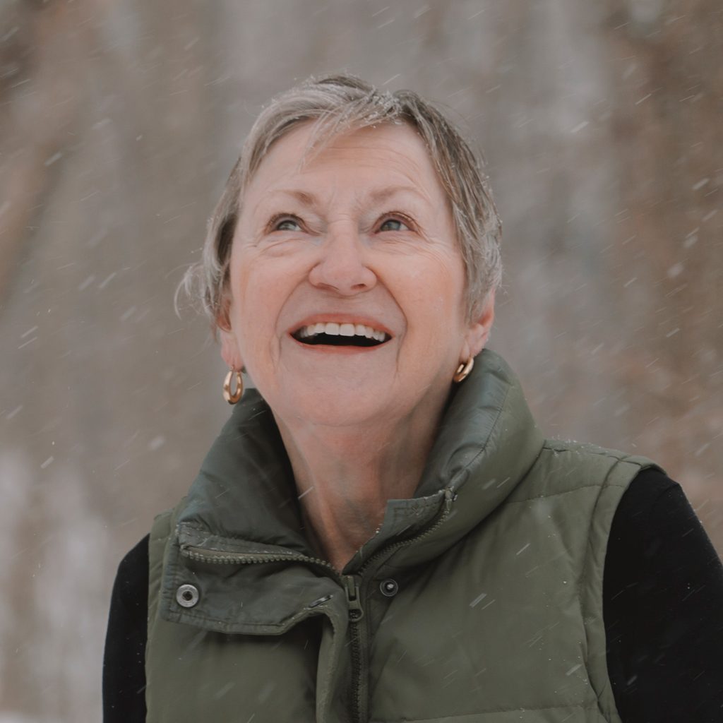 A smiling woman looks up during a snowfall.
