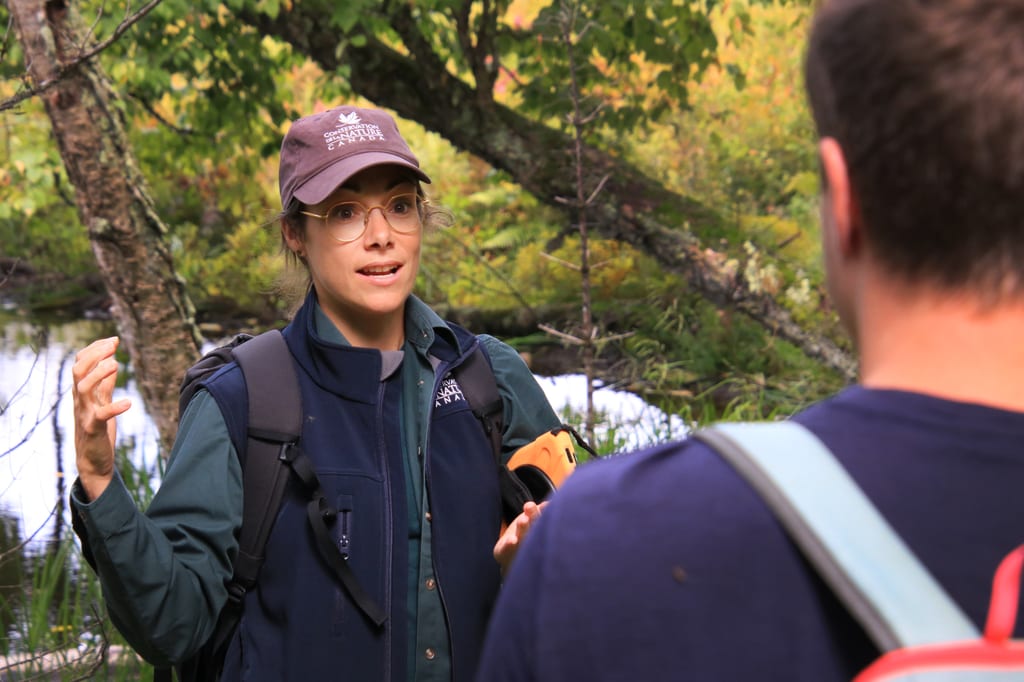 A woman in a vest and hat gestures while talking to another person in a wooded area near water.