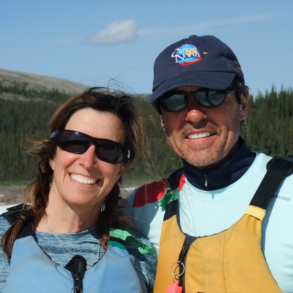 A man and a woman wearing life jackets smile in front of a forested landscape.