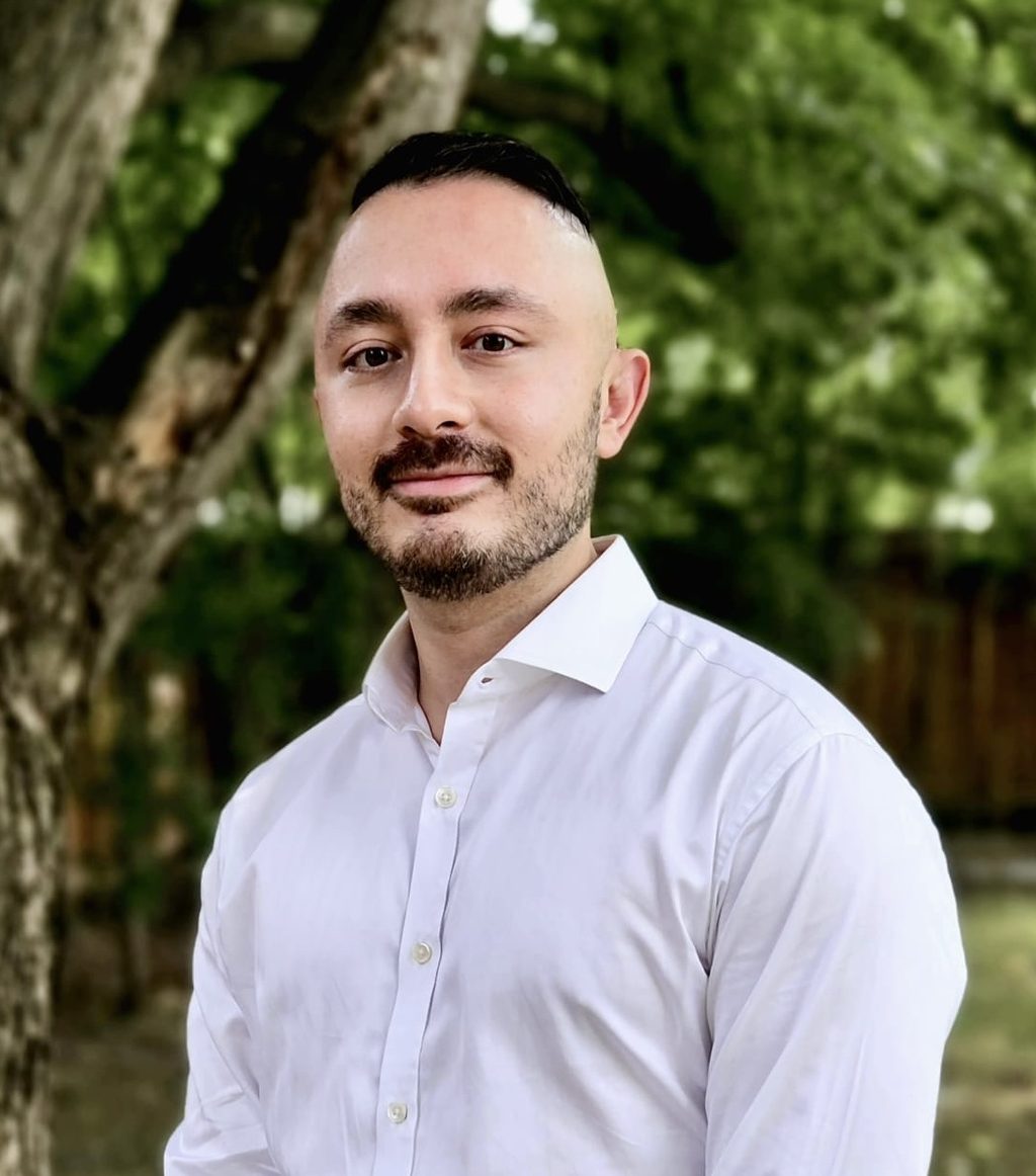 A man in a white shirt stands in front of green trees.
