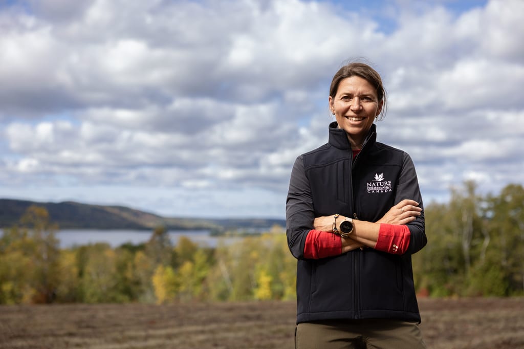 A woman wearing a Nature Canada jacket stands in front of a landscape with a lake and autumn trees.