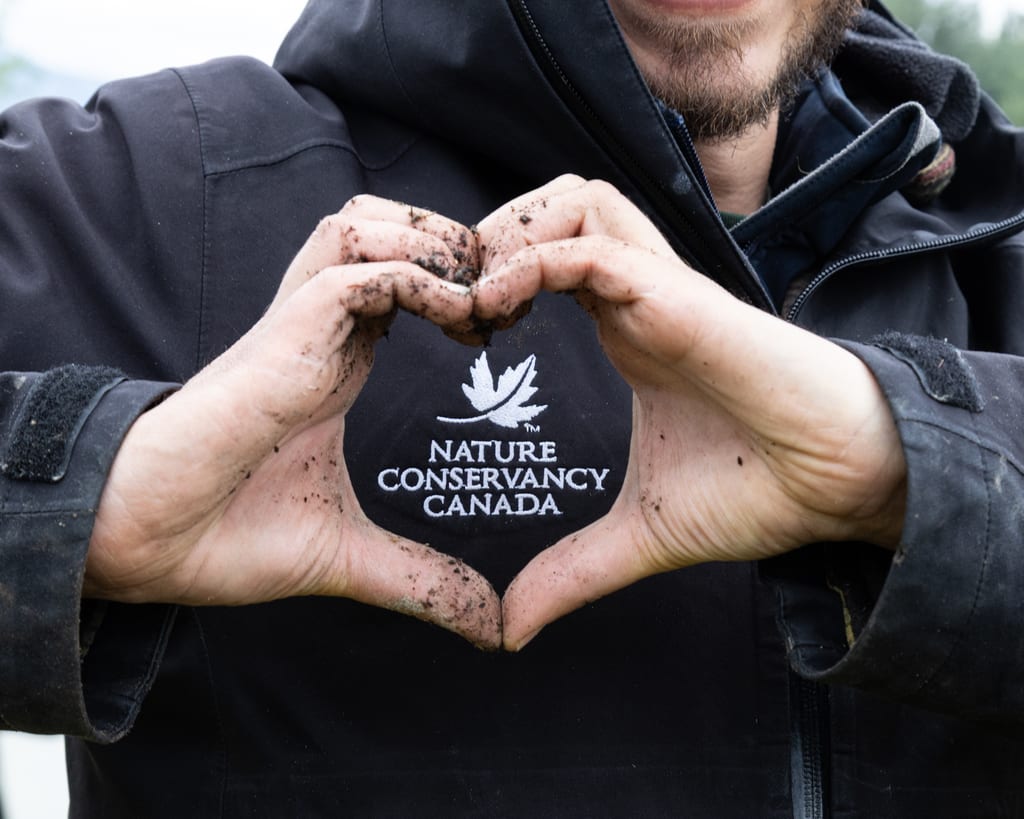 A person wearing a black jacket with the Nature Conservancy Canada logo makes a heart shape with their dirty hands.
