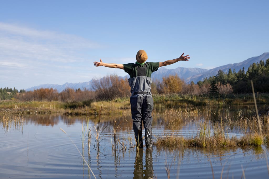 A NCC staff member in waders stands in a wetland with arms outstretched against a backdrop of mountains and trees.