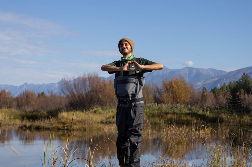 A NCC staff member in waders makes a heart shape with his hands in a wetland with mountains in the distance.