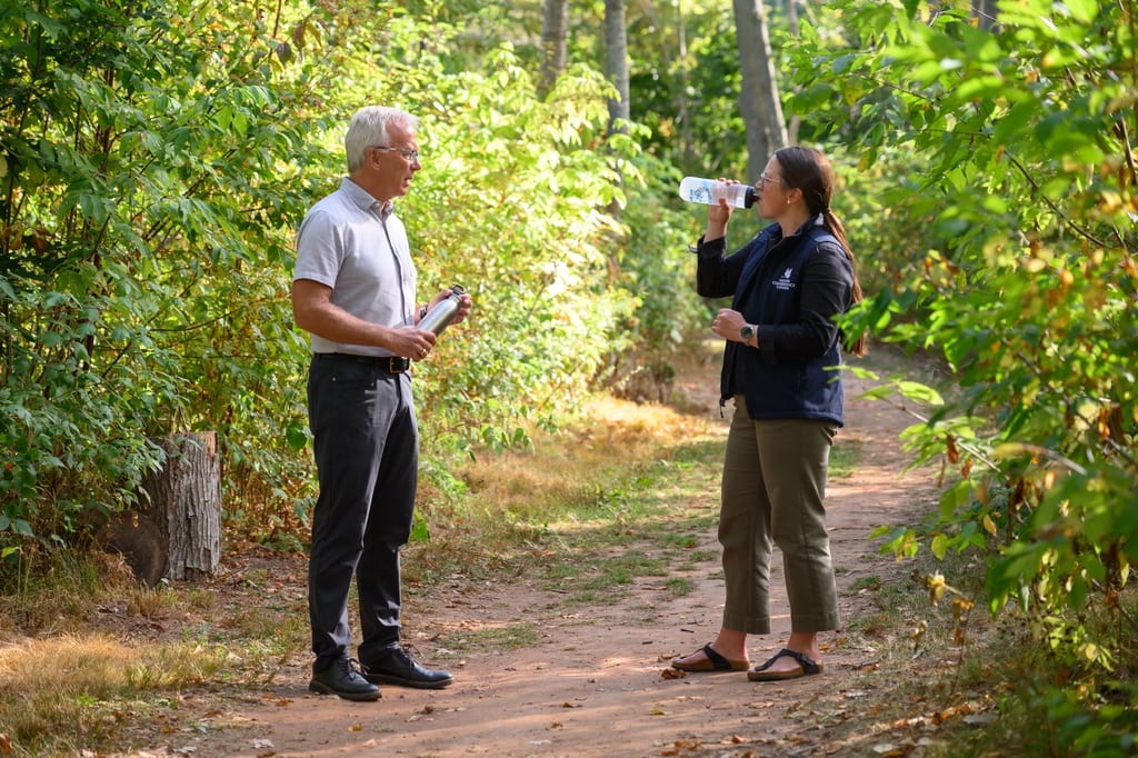 A man and a woman stand on a dirt path in a lush green forest.