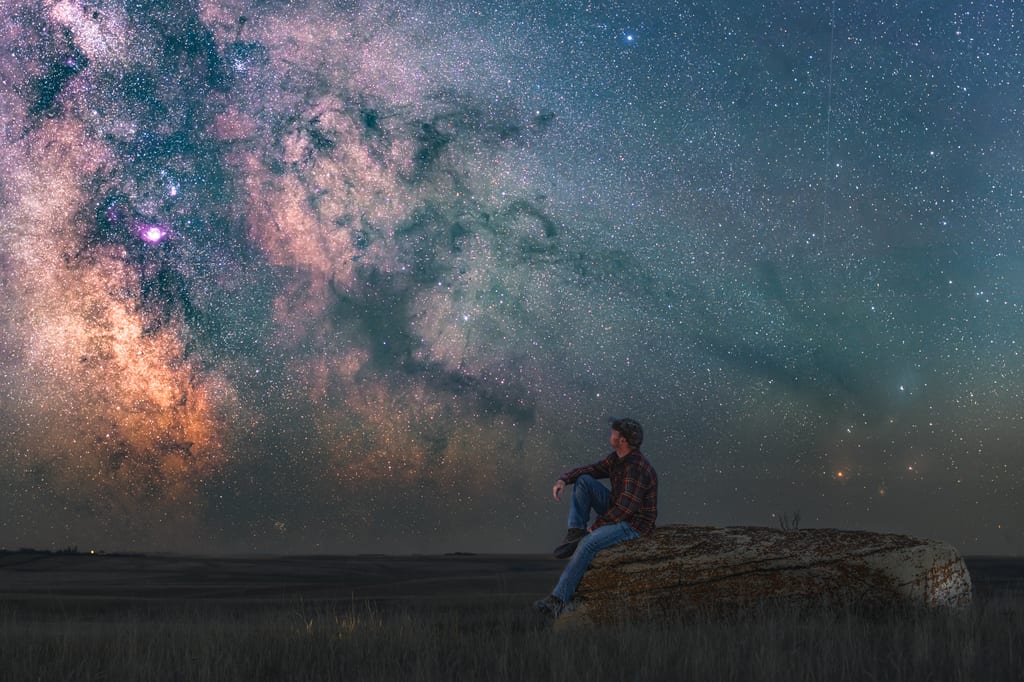 A person sits on a rock in a field under a starry night sky with the Milky Way.