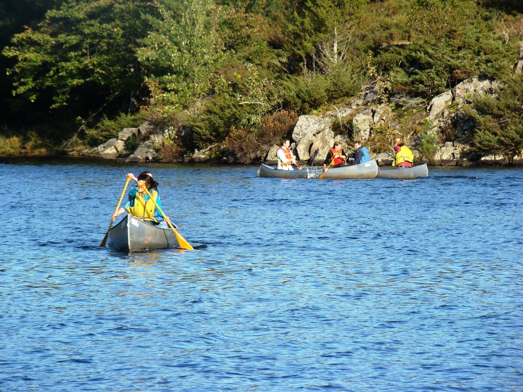 People paddle canoes on a blue lake near a rocky, forested shoreline.