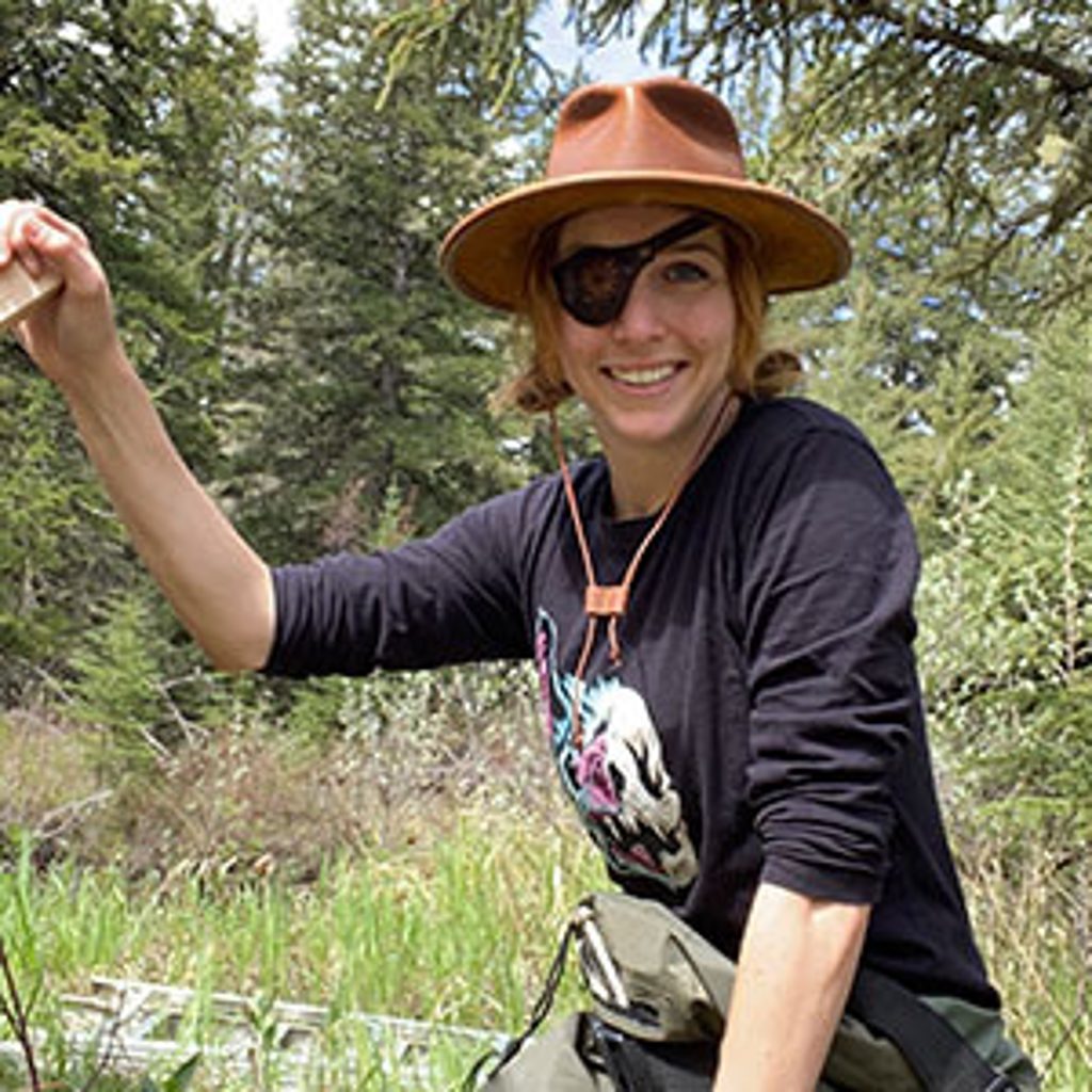 A person in a brown hat and eye patch smiles while holding an object in a wooded area.