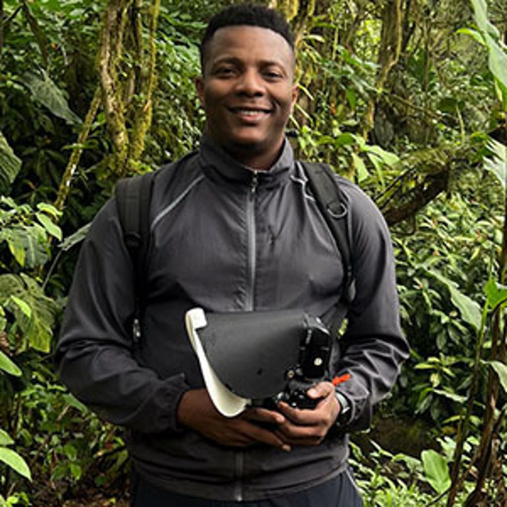 A man smiles while holding a camera in a lush forest.