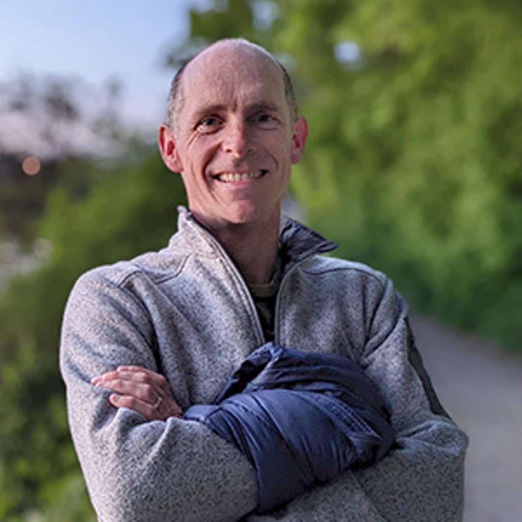 A smiling man stands with his arms crossed in front of green foliage.