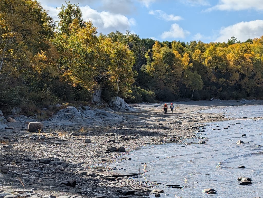 Two people walk along a rocky shoreline next to a forest with autumn foliage.