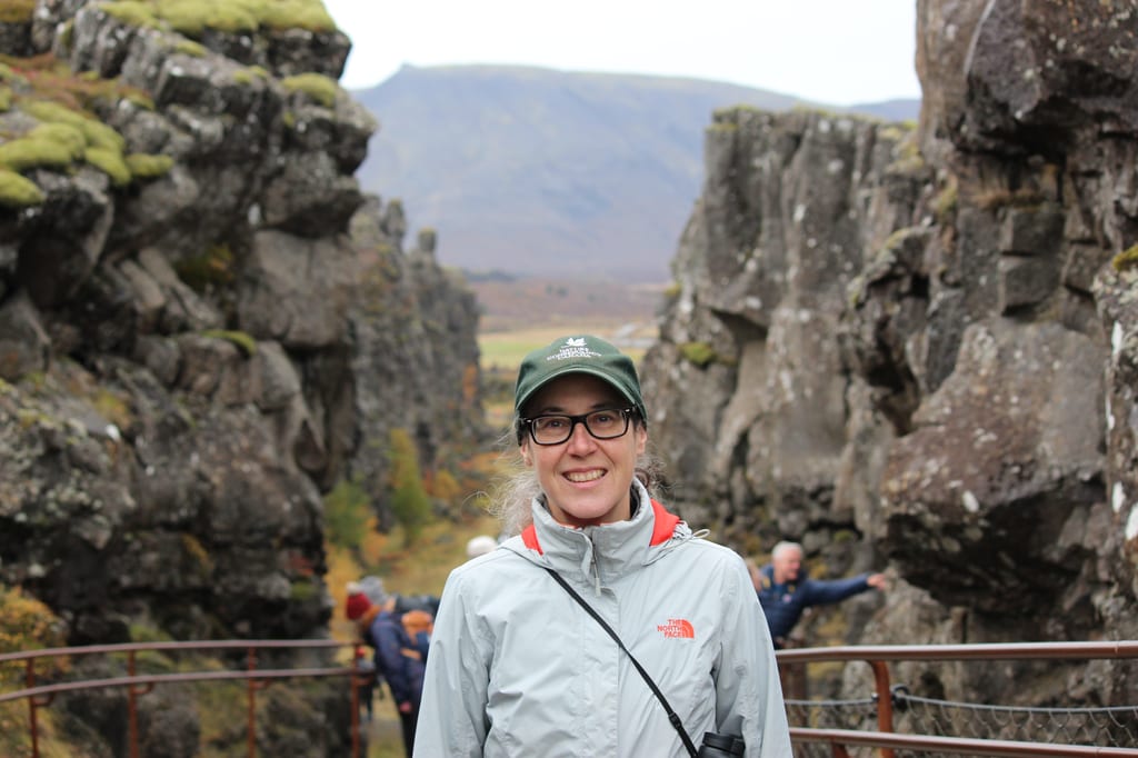 A woman stands in a rocky canyon with mossy cliffs and mountains in the distance.