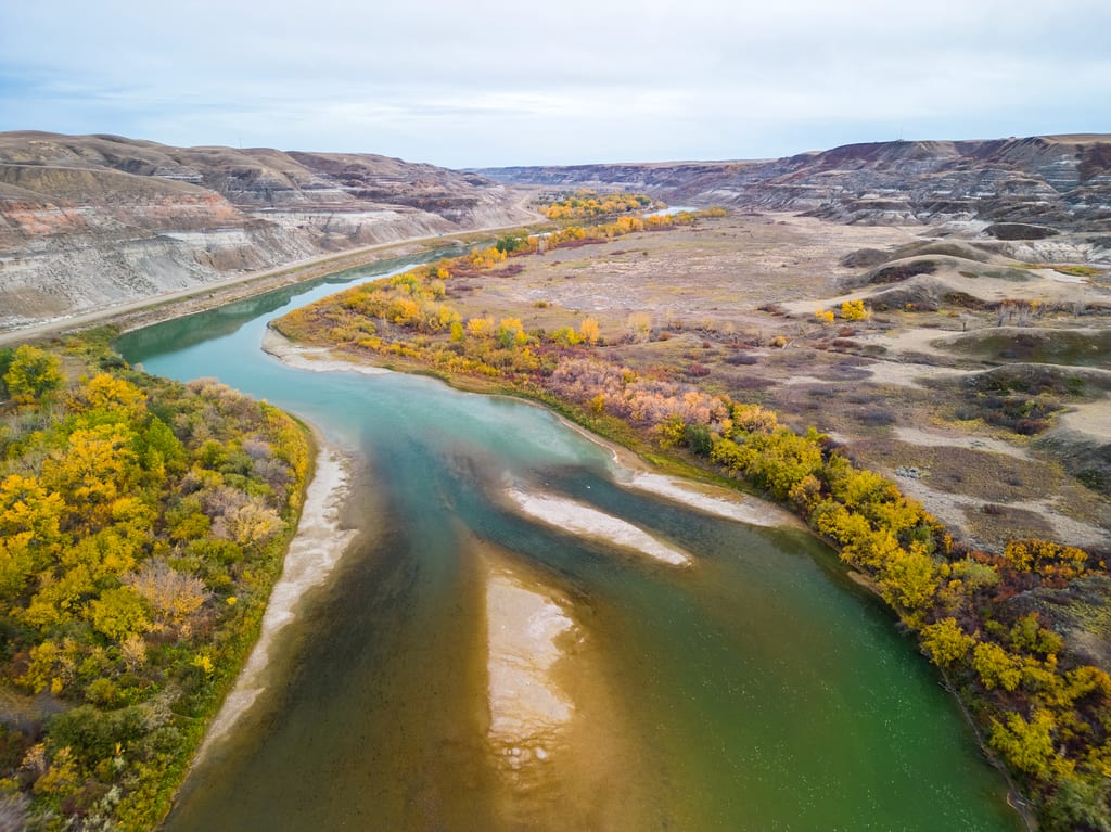 A winding river flows through a valley with autumn foliage along its banks and eroded hills in the background.