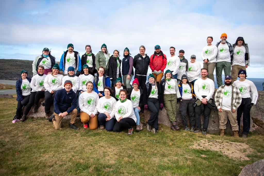 A group of people poses for a photo outdoors on a grassy hill with many wearing white NCC T-shirts.