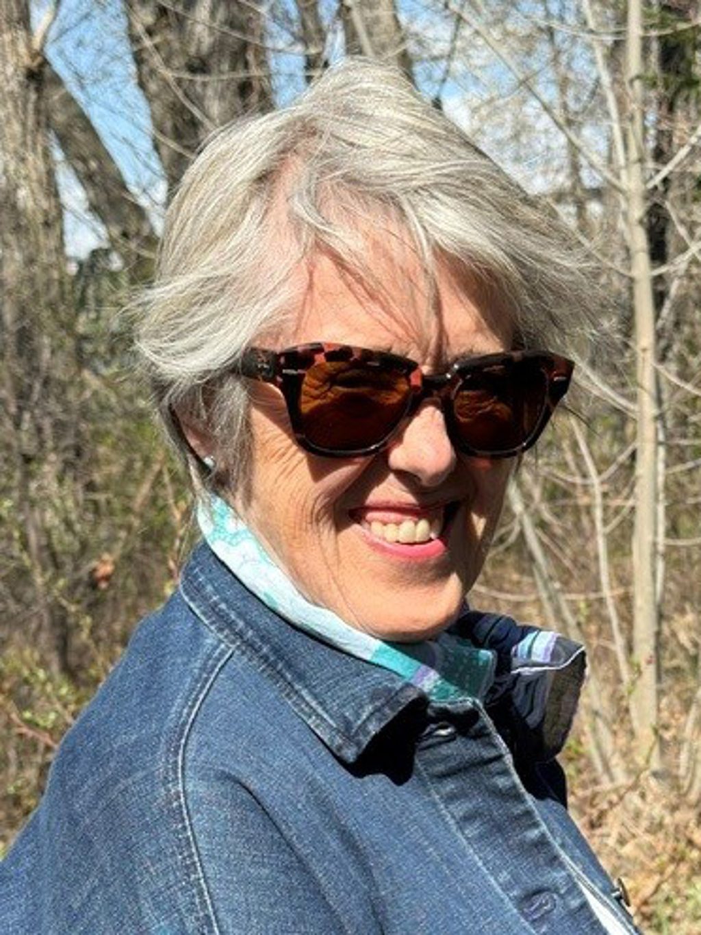 A smiling woman wearing sunglasses and a denim jacket in a wooded area.