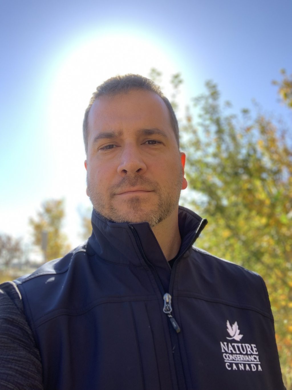 A man wearing a Nature Conservancy Canada vest stands outdoors in front of trees with autumn foliage.