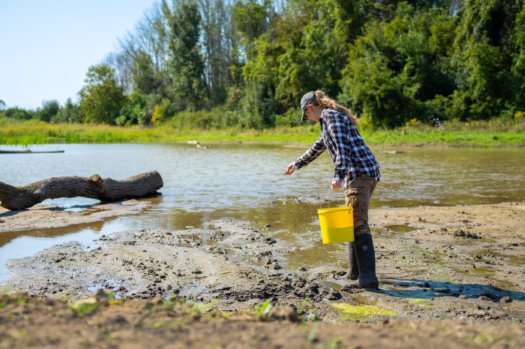A person in a plaid shirt carries a yellow bucket while walking through the mud near a pond.