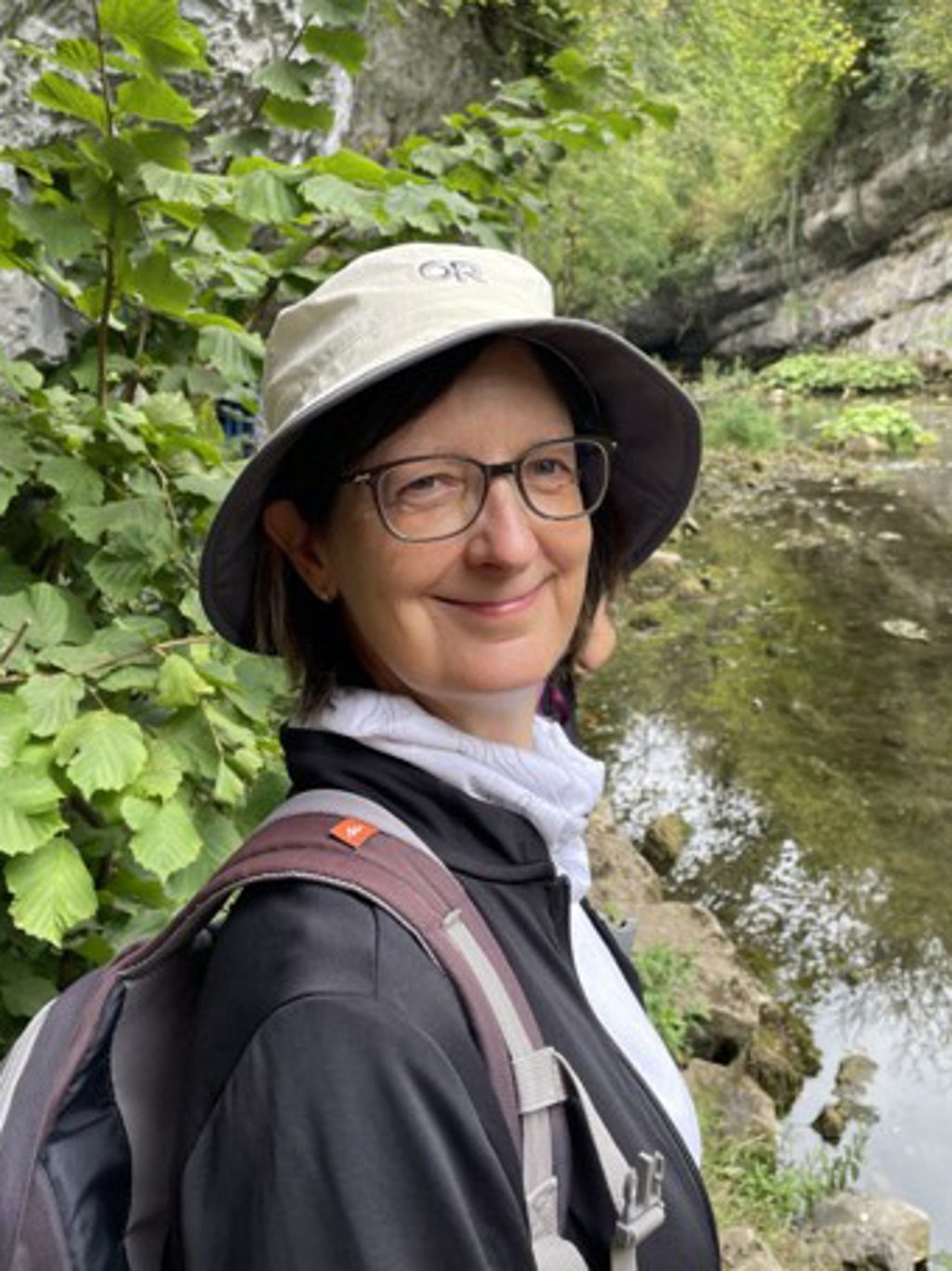 A woman wearing a hat and backpack smiles while hiking near a stream.