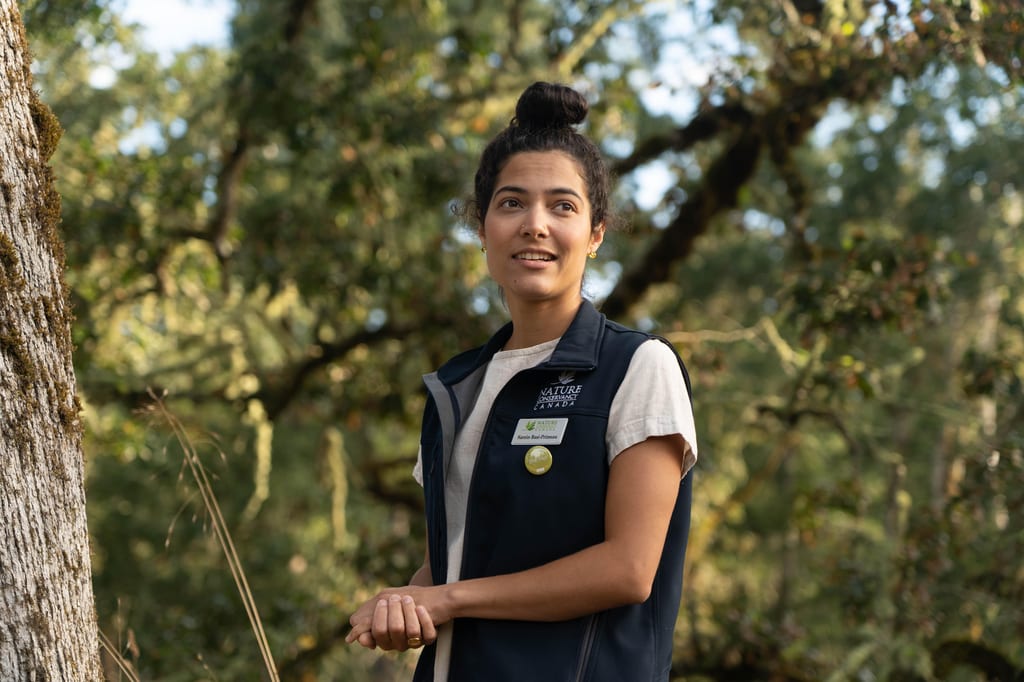 A NCC staff member wearing a Nature Conservancy of Canada vest stands in a lush green forest.
