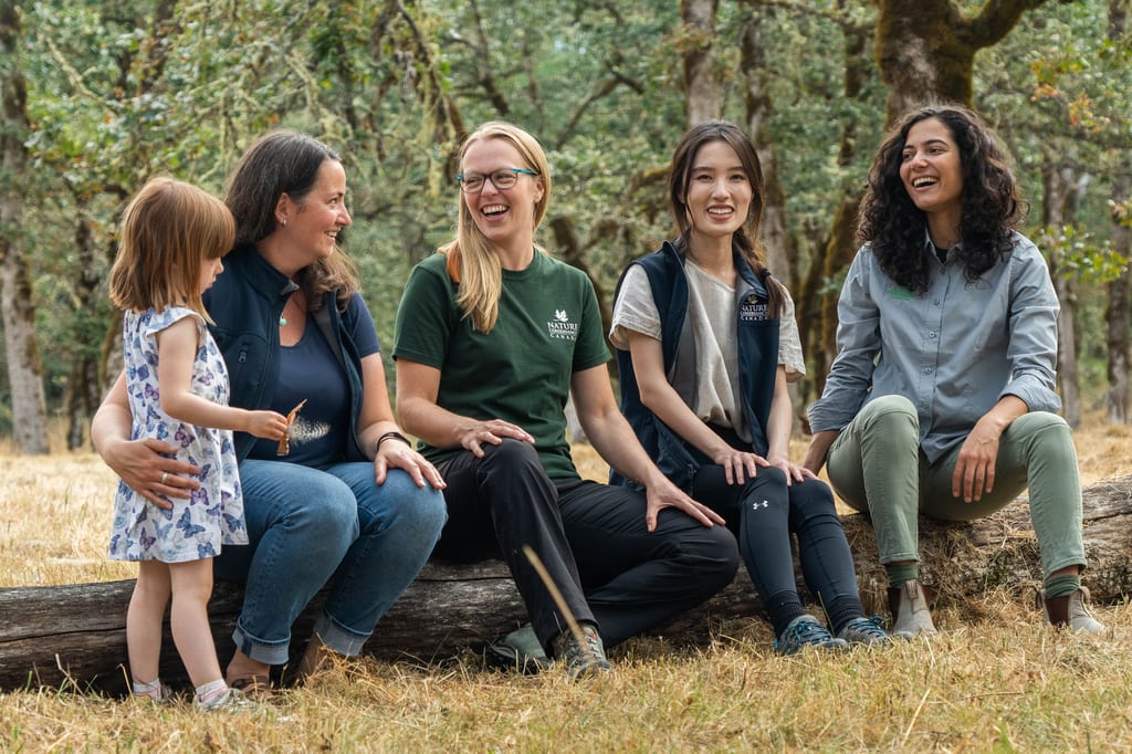 A group of people sitting on a fallen log in a wooded area.
