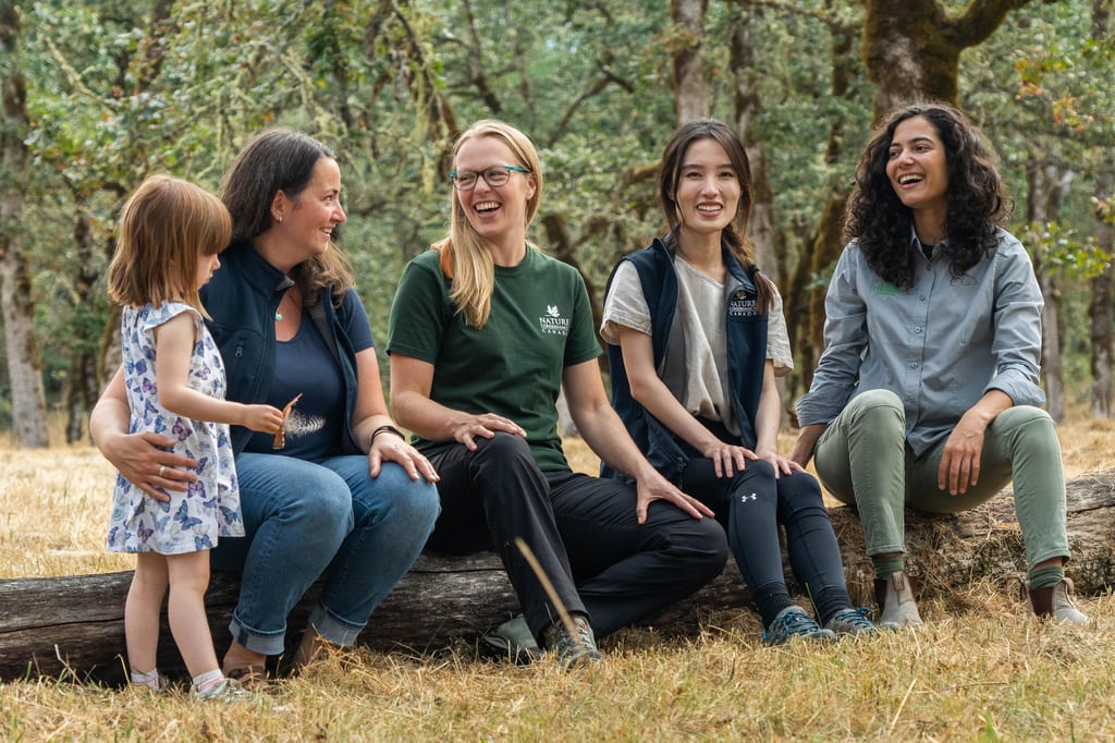 A group of people and a young child sit on a fallen log in a wooded area.