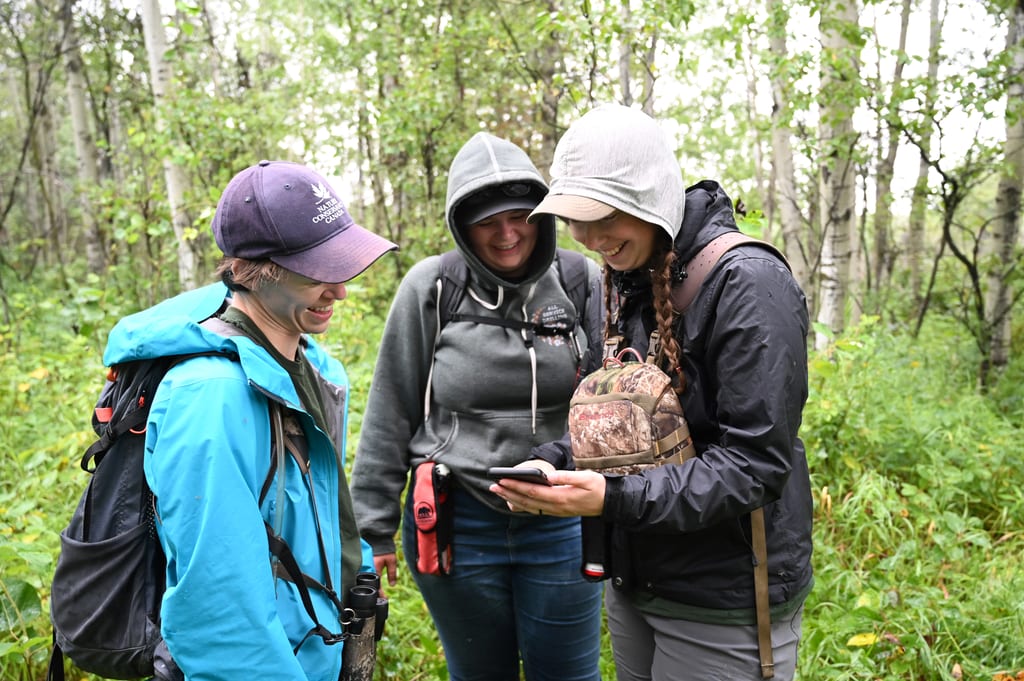 Three women look at a smartphone together in a lush green forest.