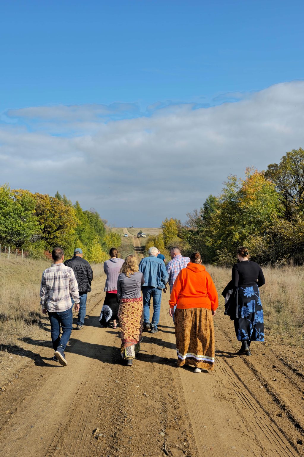 A group of people walks down a dirt road through a field with autumn trees under a blue sky.