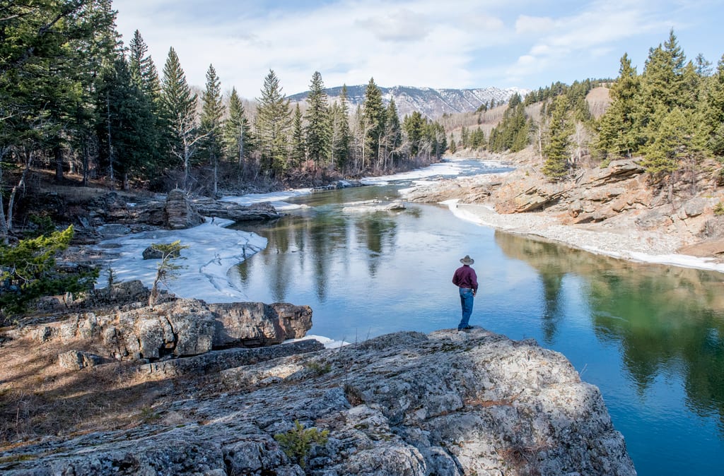 A person stands on a rocky outcrop overlooking a river flowing through a coniferous forest with ice on the banks.