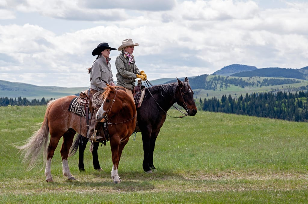 Two people ride horses through a grassy meadow with hills and forests in the background.