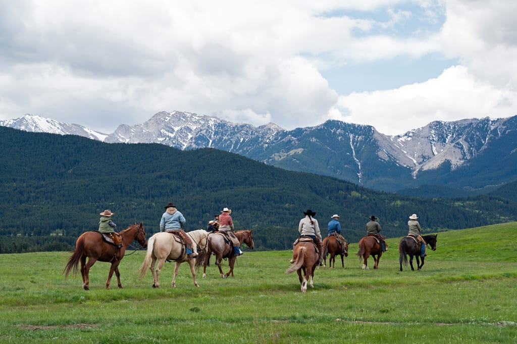 People riding horses through a green meadow with mountains and forests in the background.