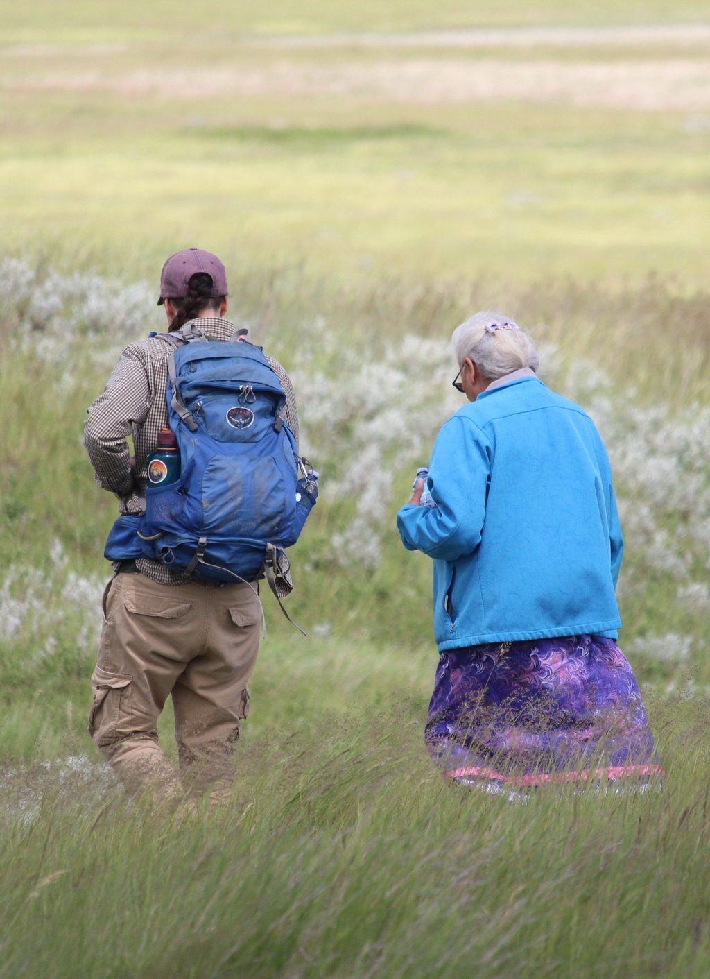 Two people walk through a grassy field, one carrying a blue backpack and the other wearing a bright blue jacket.