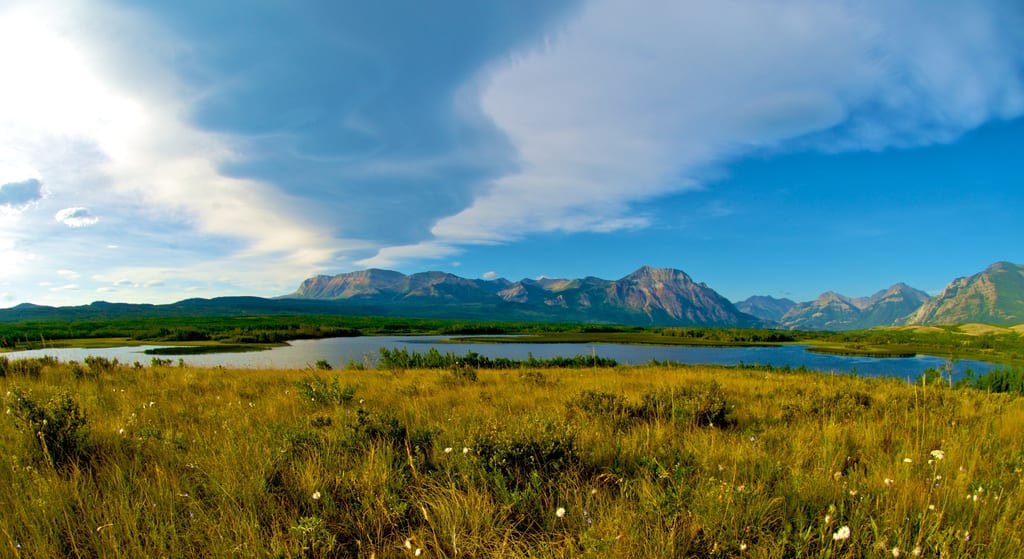 A lake is surrounded by grassy meadows with mountains in the background under a blue sky.