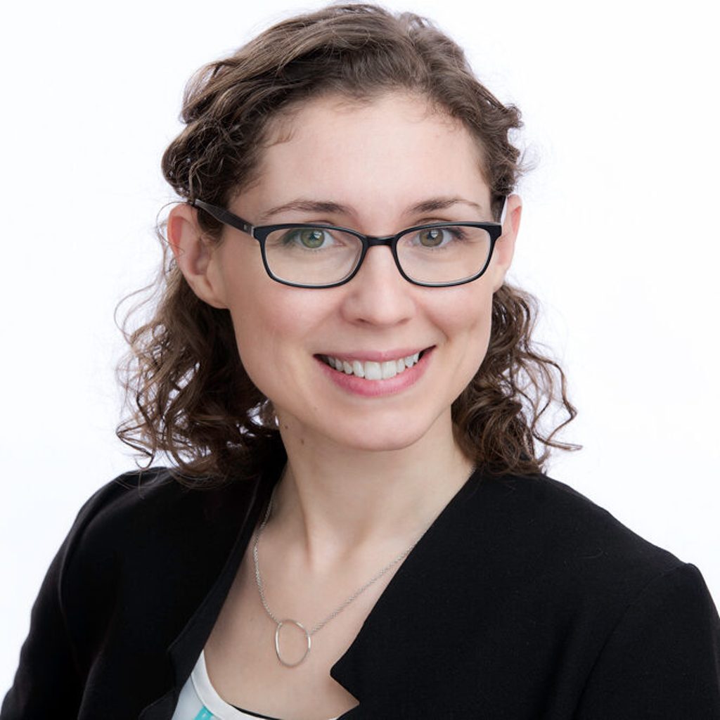 A woman with curly hair and glasses smiles against a white background.