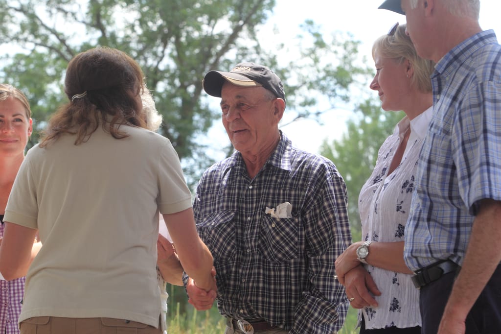 A woman shakes hands with a man wearing a baseball cap and a plaid shirt, while other people stand nearby outdoors.