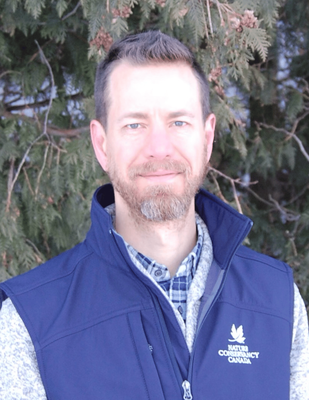 A man wearing a blue vest with the Nature Conservancy Canada logo stands in front of evergreen trees.