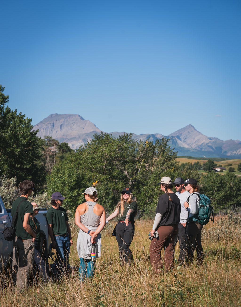 A group of people stands in a meadow with mountains in the background.