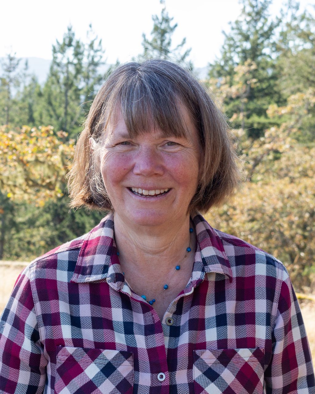 A smiling woman in a plaid shirt stands in front of a wooded landscape.