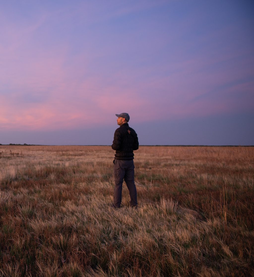 A man stands in a field of dry grass under a purple twilight sky.
